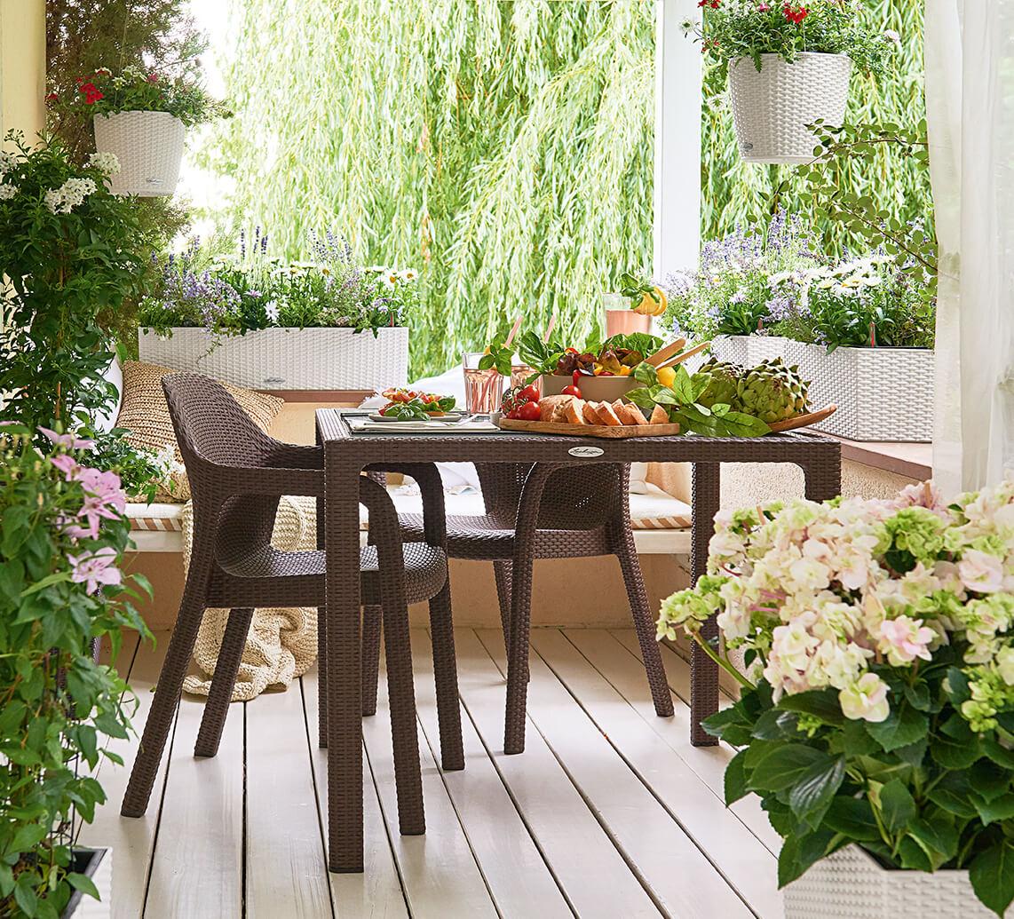 On a covered country veranda there is a beautifully set LECHUZA garden table with two chairs. It is surrounded by various LECHUZA planters in wickerwork look which are planted with various flowers e.g. hydrangeas and orleander.
