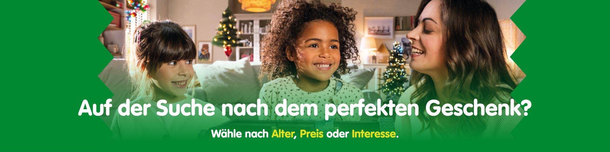 Zwei Kinder und eine Frau sitzen l&auml;chelnd vor einem weihnachtlich geschm&uuml;ckten Wohnzimmer mit Tannenbaum &ndash; perfekte Geschenkideen f&uuml;r Kinder entdecken.